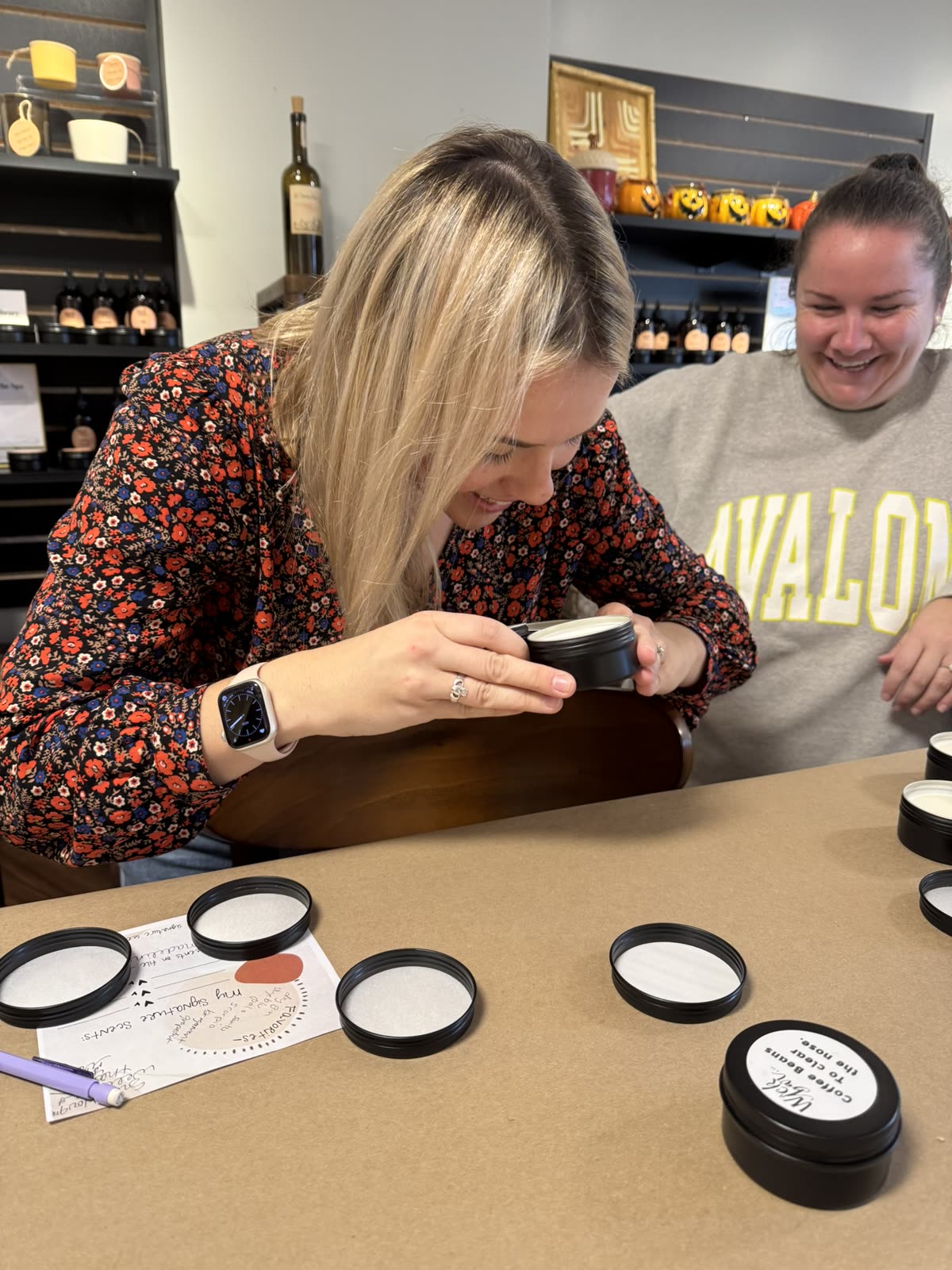 Two friends laughing as they smell fragrance tins at the Wick Bar table.