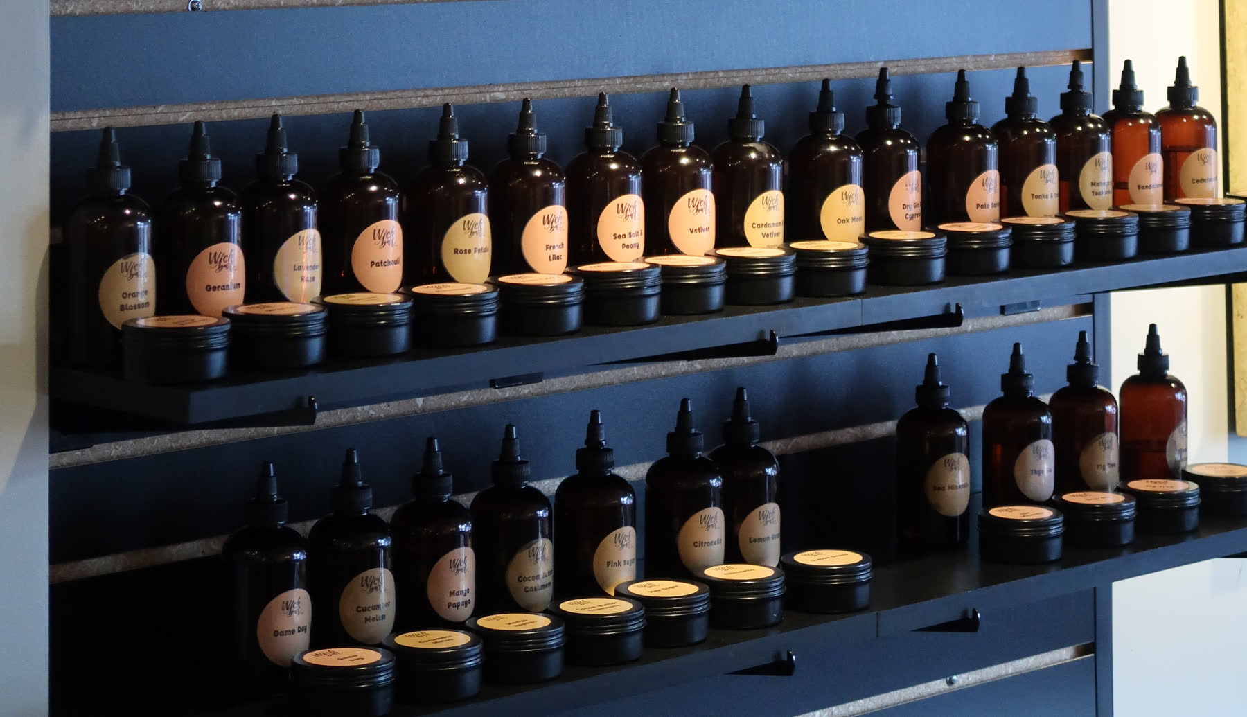 The Wick Bar fragrance wall — two rows of amber fragrance bottles on navy shelving.