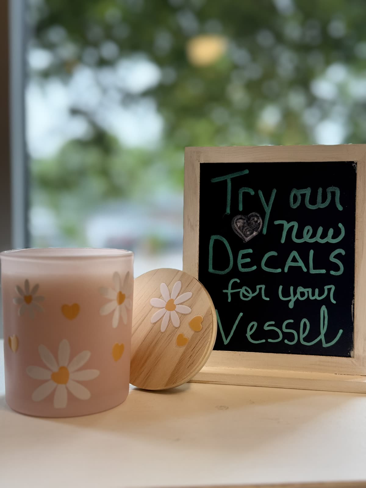 A candle with daisy decals beside a chalkboard: “Try our new decals for your vessel.”