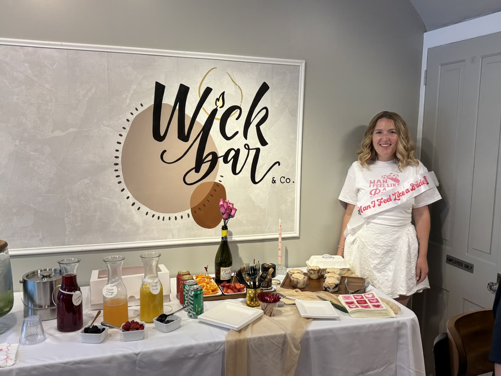 A bride in her sash beside her party spread under the Wick Bar sign.